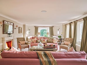 Un salon chaleureux avec une cheminée, deux canapés, une table basse agrémentée d’objets décoratifs et une vue sur la verdure à l’extérieur.