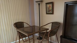 Une pièce comprenant une table à plateau en verre, deux chaises en osier, un lampadaire, un tableau encadré accroché au mur et un meuble TV de couleur sombre.
