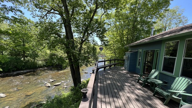 Une terrasse en bois, agrémentée de chaises, qui offre une vue sur une rivière et un bâtiment aux volets verts.
