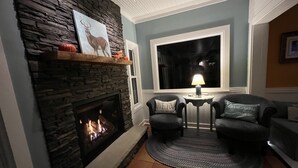 Un salon chaleureux avec une cheminée en pierre, une grande fenêtre et deux fauteuils agrémentés de coussins.