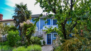 Ein zweistöckiges Haus mit blauer Tür und weißen Fensterläden, umgeben von üppiger Vegetation und einem Kiesweg, der zum Eingang führt.