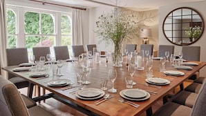Une salle à manger avec une longue table en bois dressée pour un repas, entourée de chaises, et agrémentée d’un vase de fleurs et d’un miroir rond accroché au mur.