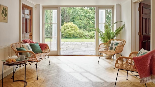 Une pièce spacieuse avec deux fauteuils en osier, une table en bois sur laquelle se trouve un plateau de croissants, et une grande porte vitrée donnant sur un jardin extérieur.