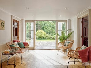 A spacious room with two wicker chairs, a wooden table with a tray of croissants, and a large glass door leading to an outdoor garden. - The Lindens, spacious Dorset retreat for a coastal break (East Lulworth)