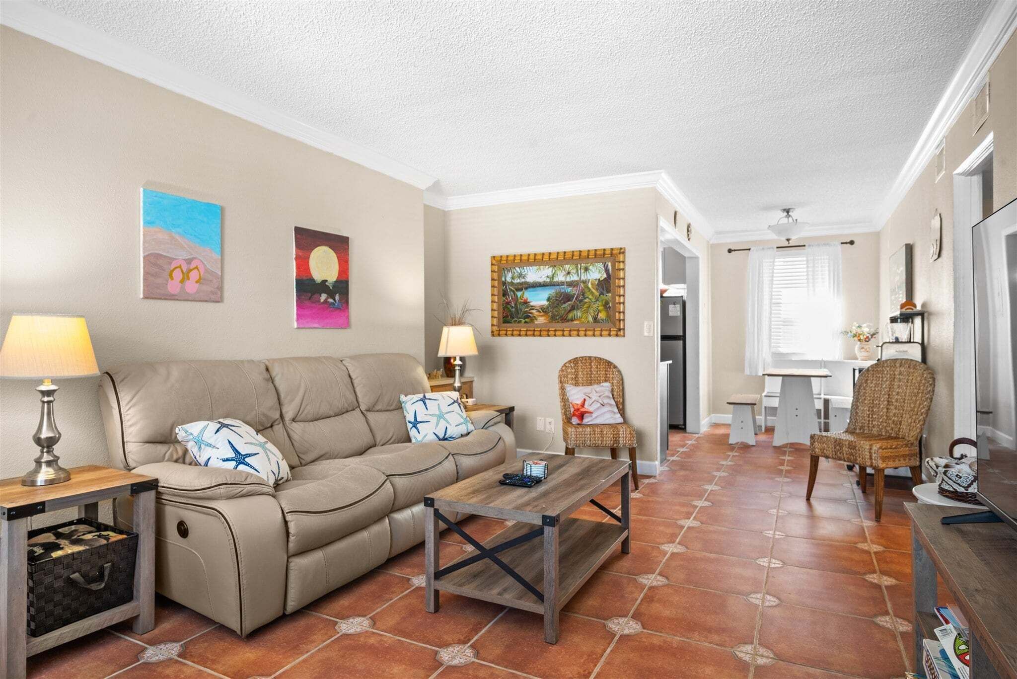 A living room with a sofa, coffee table, and two chairs. There are paintings on the walls and a dining area in the background.