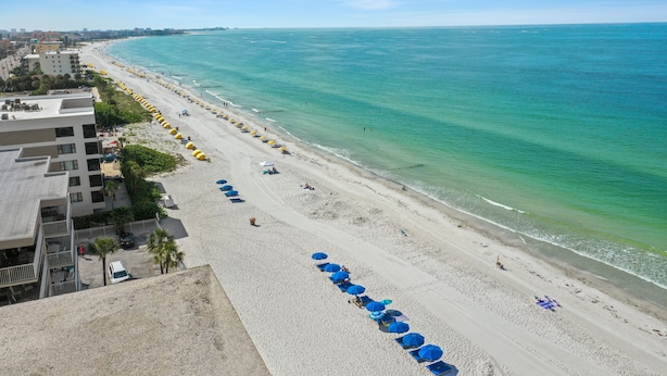 Ein Strand mit blauen Sonnenschirmen und Liegestühlen, eine sandige Küstenlinie und Gebäude im Hintergrund.
