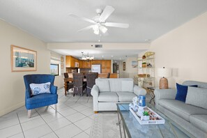 A spacious living room with a ceiling fan, a dining area with chairs, and a kitchen in the background.