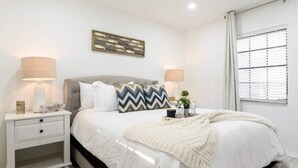 A bedroom with a bed, two bedside tables, a nightstand, a wall-mounted artwork, a window with blinds, and a lamp on each bedside table. - Beautiful Waterfront Condo on Canal with Pool & Walk to Beach! Boat Watch! 1b/1b (Pompano Beach)