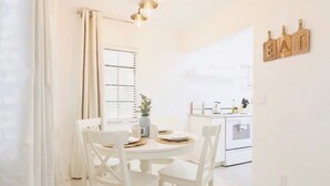 A bright, white dining area with a round table and chairs, a window with blinds, and a kitchen in the background. - Beautiful Waterfront Condo on Canal with Pool & Walk to Beach! Boat Watch! 1b/1b (Pompano Beach)