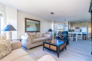 A spacious living room with a sofa, coffee table, and a dining area with chairs and a kitchen in the background. - Cheerful & Bright Spacious End Unit - Oceanfront, Porch w/ View,  Pool (Hilton Head Island)