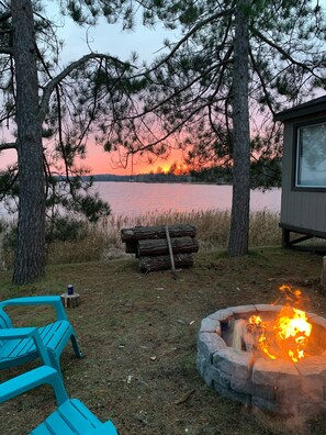 Una cabaña junto al lago con un pozo de fuego, asientos exteriores y vistas a la puesta de sol sobre el agua.