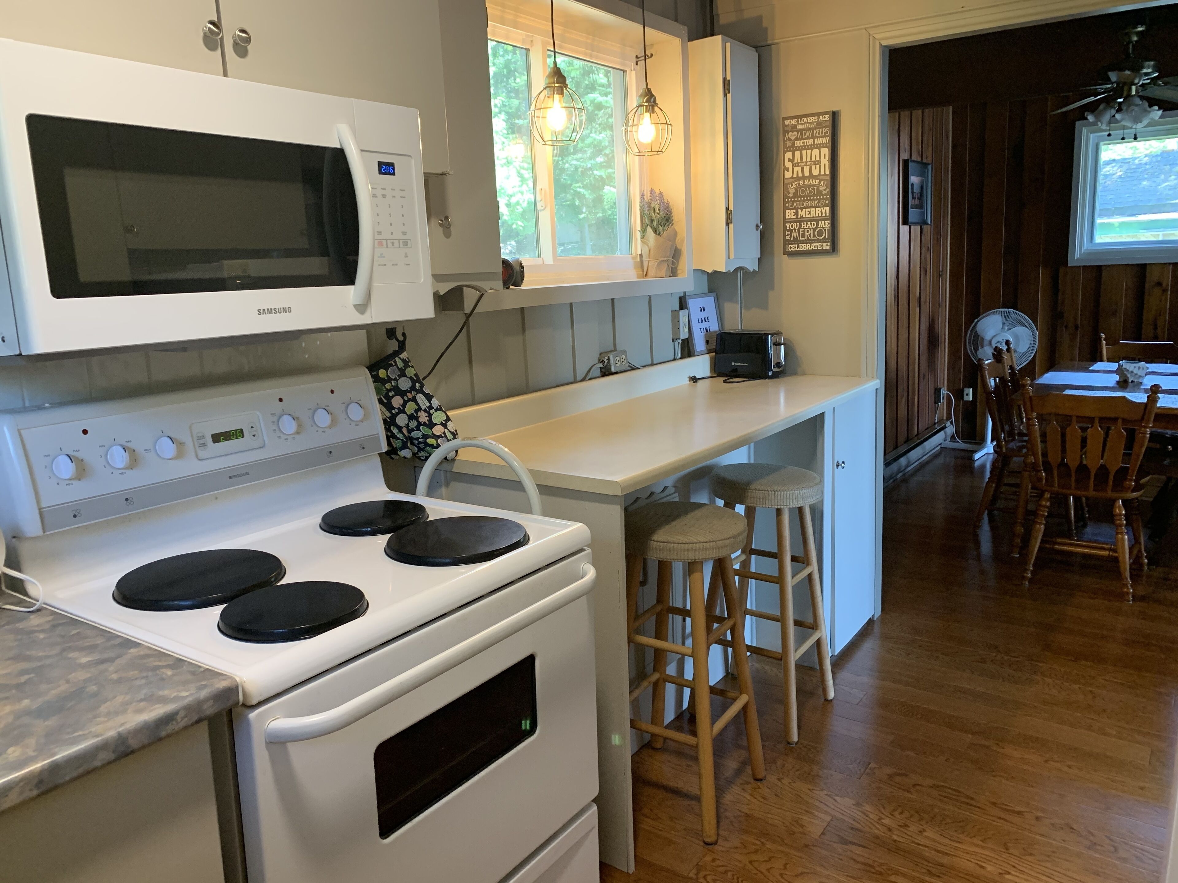 Una cucina moderna con un piano cottura bianco, un forno a microonde e un lavello. Sono presenti sgabelli di legno e un tavolo da pranzo con sedie.
