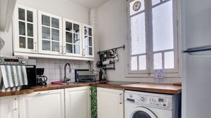 Cocina compacta con armarios blancos, lavadora integrada, ventana con reloj y encimera con diversos objetos.
