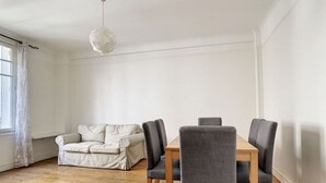 Una habitación con piso de madera, un sofá blanco, una mesa de comedor con cuatro sillas y una lámpara colgante.