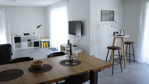 A modern living room with a wooden dining table, a TV mounted on the wall, and a coffee-themed wall decoration. - die Fewo im Herzen der Pfalz (Lug)