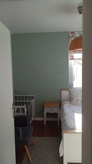 Ein Schlafzimmer mit einem Babybett, einem Nachttisch, einem Bett mit floralem Überwurf und einem Fenster mit einem Gewebeschirm.