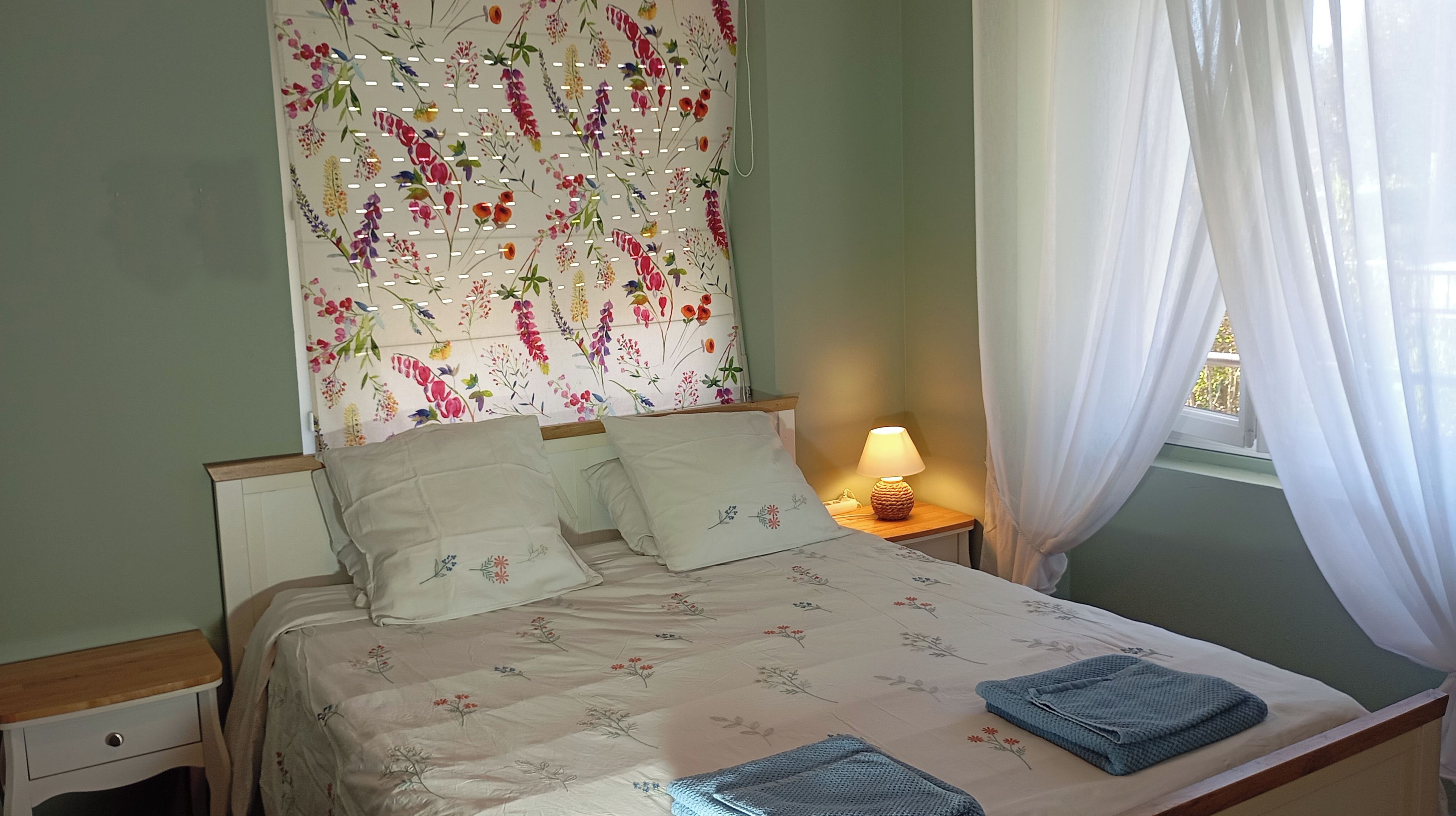 Un lit avec une literie à motifs floraux, une lampe de chevet et une fenêtre ornée de rideaux légers.