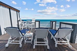 Una terraza con sillones de descanso y una mesa, con vista a una playa y un cielo despejado.