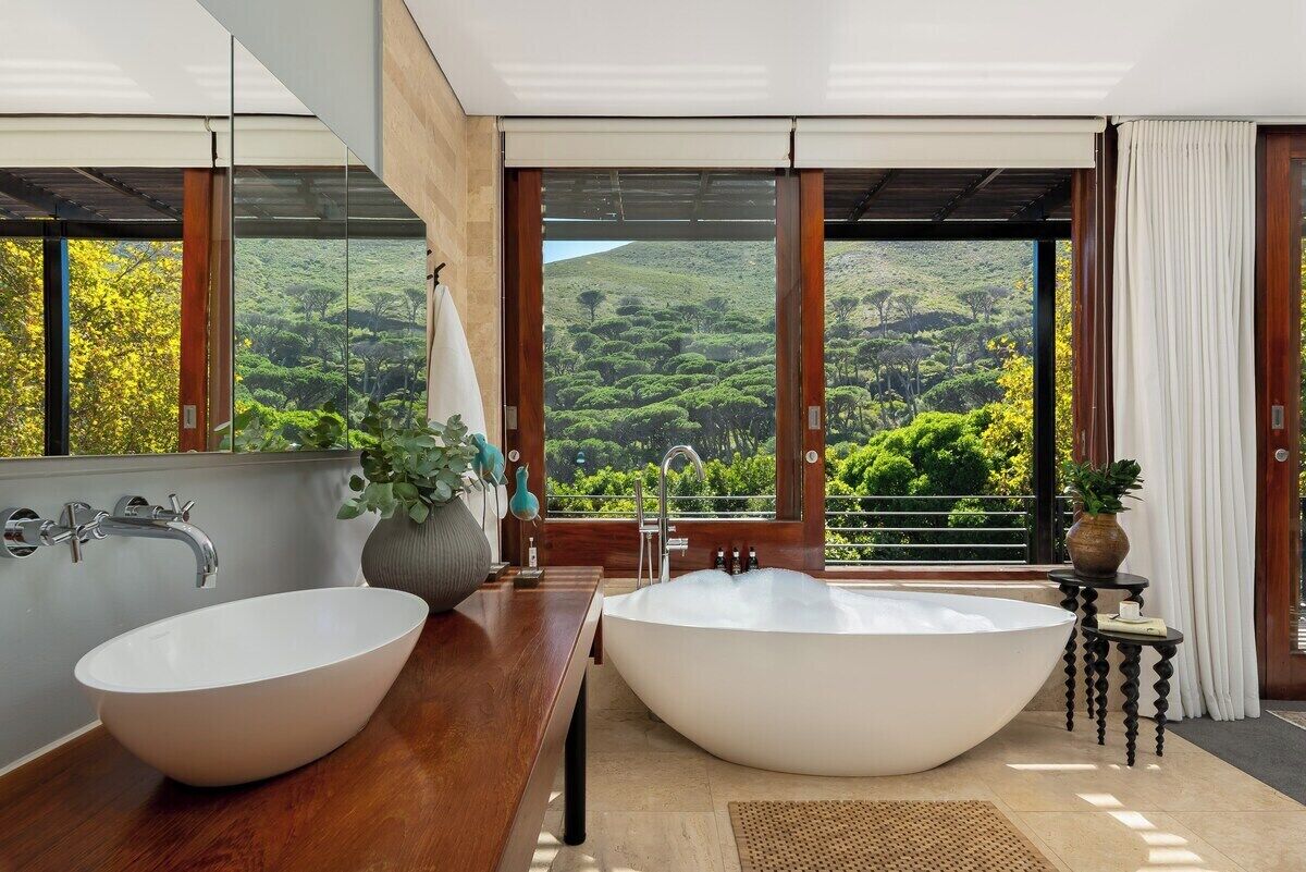 Un baño con una bañera grande y un lavamanos doble, ambos con lavabos blancos, un mueble de tocador de madera y vistas a un paisaje exuberante a través de amplios ventanales.