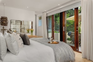 A modern bedroom with a large bed, a coffee table, and a view of the garden through a sliding glass door.