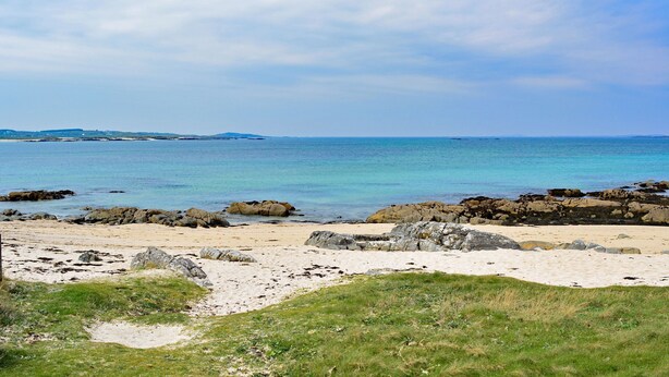 Ein Strand mit klarem, blauem Wasser, felsigem Ufer und in der Ferne Landformen.