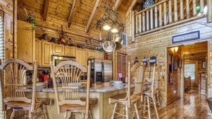 A wooden cabin interior with a kitchen area featuring a bar with wooden chairs, a stainless steel refrigerator, and a hanging light fixture.