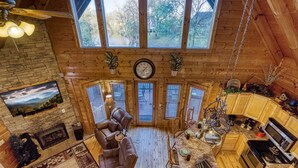 A cozy cabin interior with a fireplace, wooden furniture, a dining area, and large windows offering a view of the outdoors.