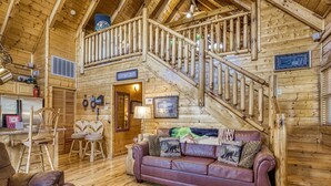 A wooden cabin interior with a staircase, a living room with a sofa and chairs, and a kitchen area.