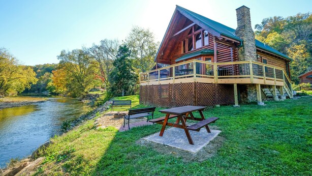 A log cabin with a deck overlooking a river, picnic table, and benches.