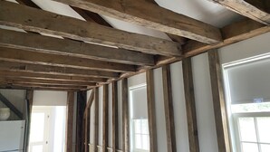 A room with exposed wooden beams and white wainscoting, large windows, and a door leading to another room.