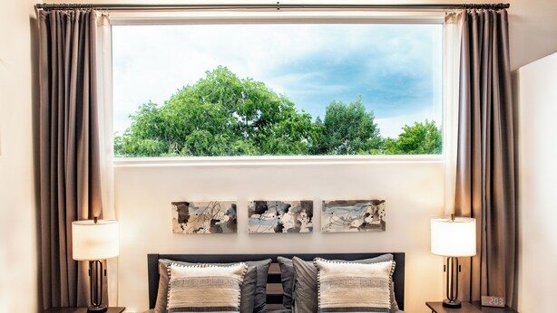 A bedroom with a large window, a bed with striped pillows, two bedside tables with lamps, and abstract wall art. - Spacious & unique railyard loft w/panoramic balcony views (Santa Fe)