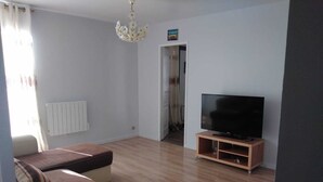 A living room with a sofa, a TV on a wooden stand, a radiator, and a chandelier. - Large F3 apartment with balcony (Pierrelaye)