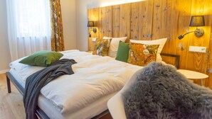 A bedroom with a wooden headboard, two bedside tables with lamps, a bed with white bedding and multiple pillows, and a window with sheer curtains.