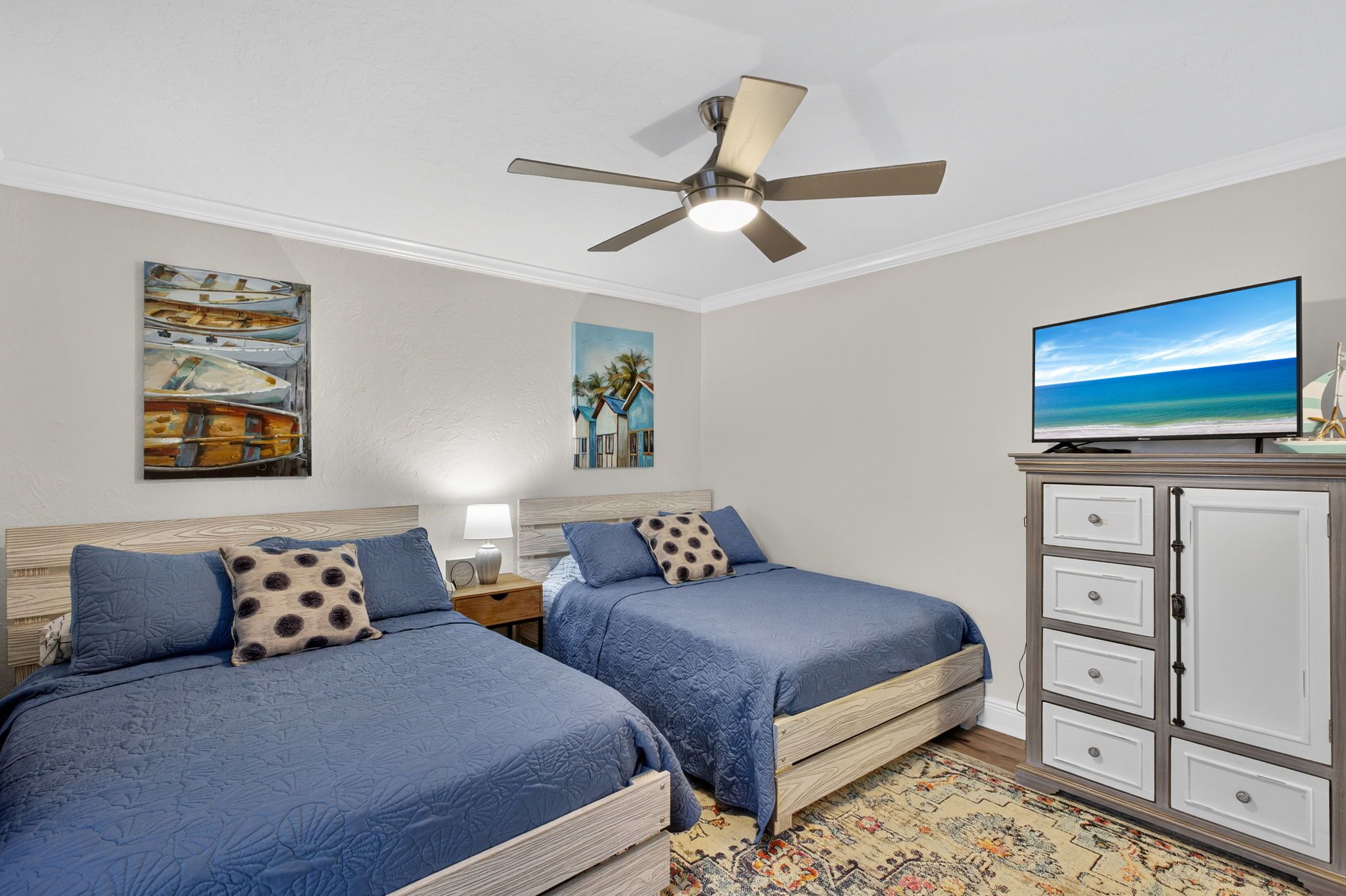 A bedroom with two beds, a ceiling fan, a TV, and artwork on the walls.