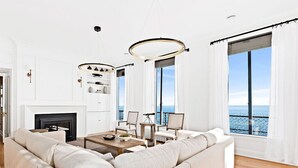 Un salón amplio con un sofá grande, una chimenea y vistas al mar a través de ventanas enormes.