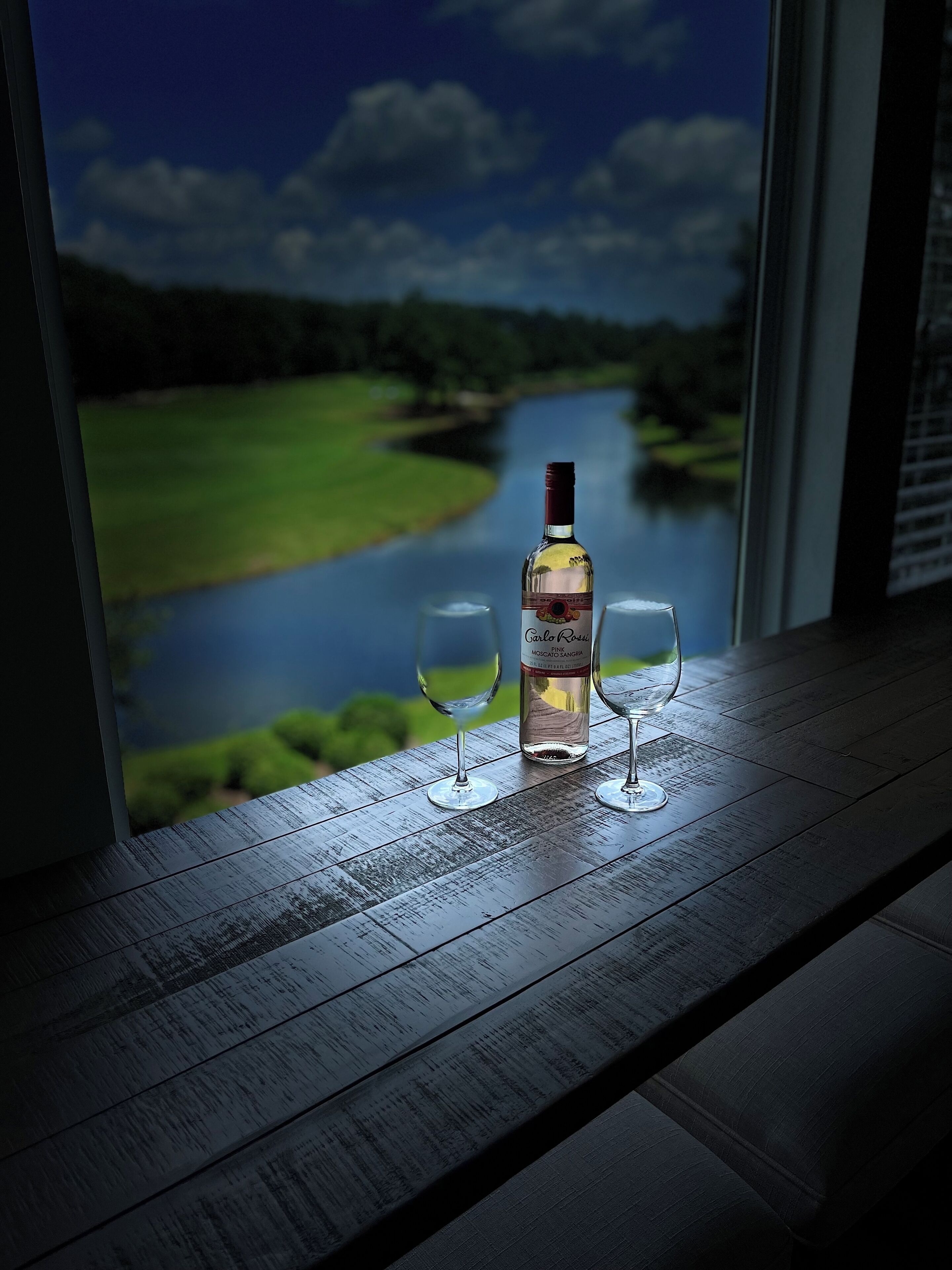 A bottle of wine and two glasses on a wooden table by a window with a view of a river and greenery.