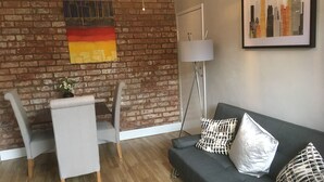 A modern living room with a brick wall, a dining area with a table and chairs, a grey sofa with patterned cushions, and a cityscape painting. - One Bedroom Maisonette with Terrace, Kidderminster (Kidderminster)