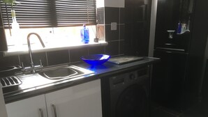 A compact kitchen with a sink, a dish rack, a washing machine, and a window with blinds. - One Bedroom Maisonette with Terrace, Kidderminster (Kidderminster)