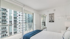 Une chambre d’hôtel moderne avec une grande fenêtre, un lit recouvert d’une couverture bleue, un miroir et un tableau représentant la Tour Eiffel accroché au mur.