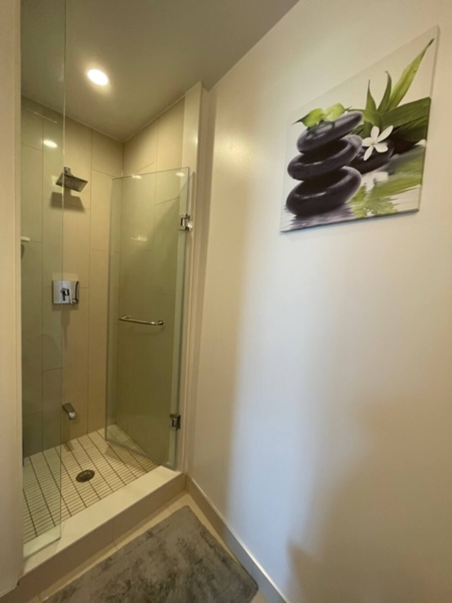 Ducha con mampara de vidrio, panel de control, piso de baldosas e imagen de piedras y flores en la pared.