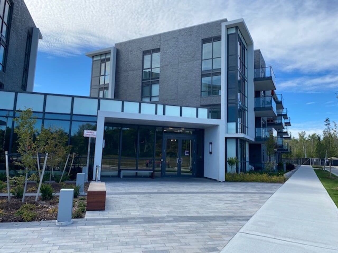 A modern building with a glass entrance, a covered walkway, and a landscaped area with trees and shrubs.