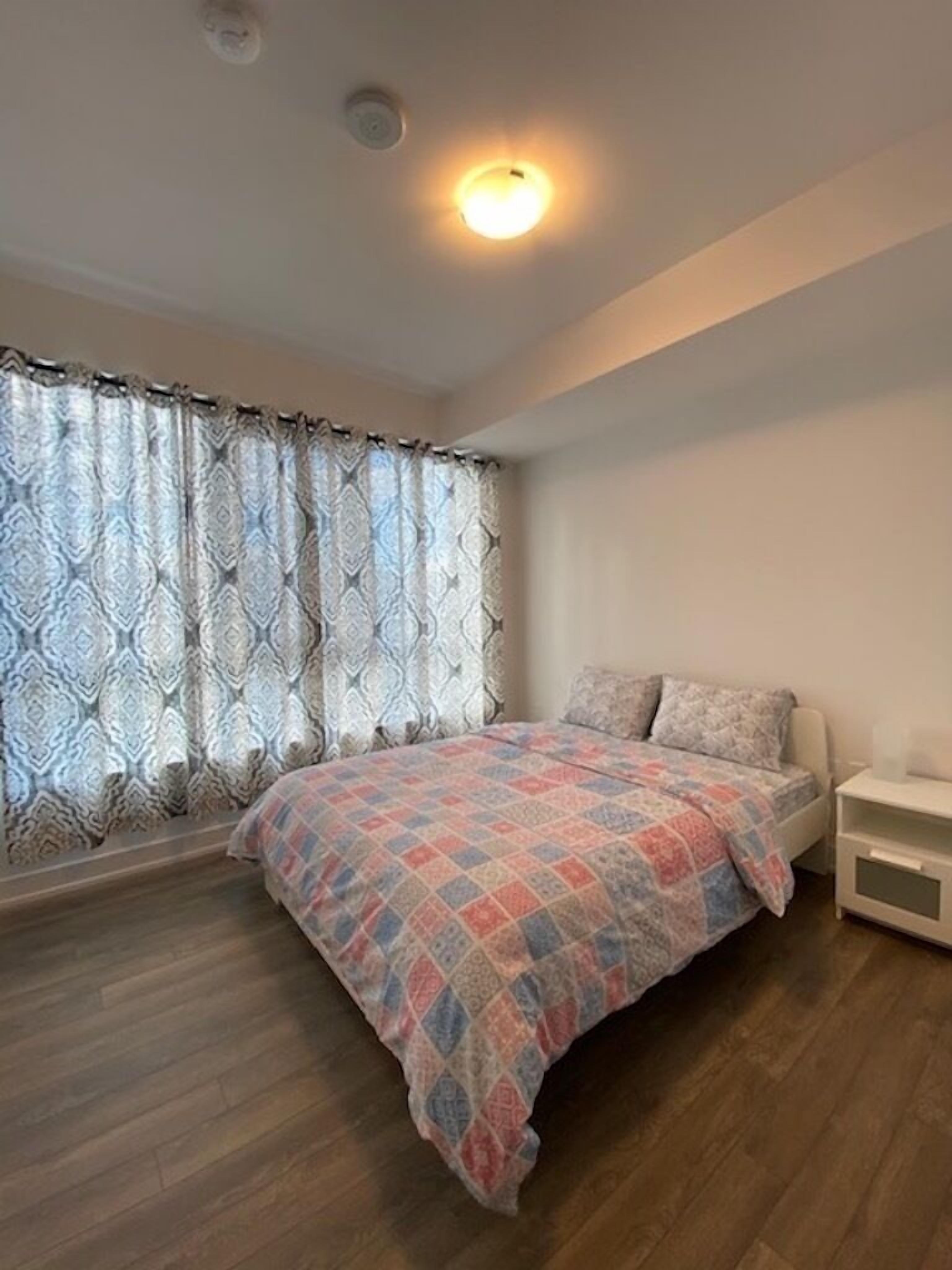 A bedroom with a bed, a nightstand, a window with patterned curtains, and a ceiling light.