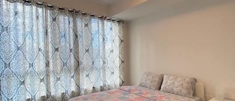 A bedroom with a bed, a nightstand, a window with patterned curtains, and a ceiling light.