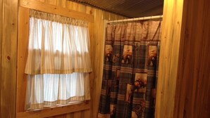 Interior de una cabaña de madera con una ventana que tiene una cortina blanca y una cortina de ducha con estampado de oso.
