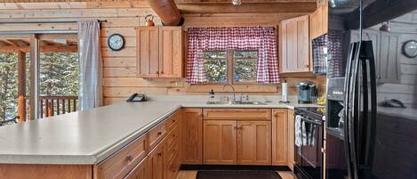 A wooden cabin kitchen with a large island, wooden cabinets, a black refrigerator, and a window with checkered curtains.