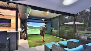 Ein Golf-Simulationsraum mit einem großen Bildschirm, einer Golfausrüstungstasche und einer Sitzecke mit blauen Kissen.