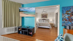 Ein Schlafzimmer mit Bett, Fernseher, Deckenventilator, einem Gemälde und Blick auf einen Strand.