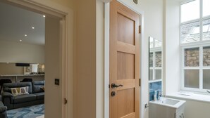 A room with a wooden door, a window with stone framing, a white sink, and a grey sofa with a cushion. - 23 Tanners Yard - Modern studio-style apartment in the heart of Kendal (Dog-friendly) (Kendal)