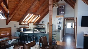 Un loft espacioso con techo alto, vigas de madera, una cocina con taburetes de bar y una zona de comedor con banco y sillas.