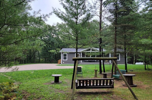 Um parquinho com balanços e mesas, cercado por árvores e uma casa ao fundo.